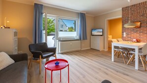 A modern living room with a sofa, a red table, a dining area with a white table and chairs, a TV, and a view of the outside through a large window. - Apartment 1 "Starfish" (ID 292) (Room category) - Apartment 1 "Starfish" (ID 292) (Sankt Peter-Ording)