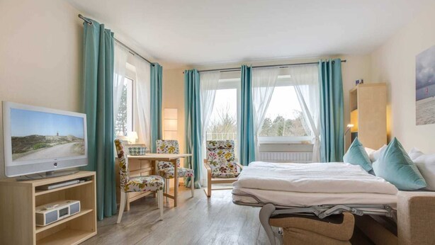 Et hotelværelse med seng, skrivebord med stol, fjernsyn og et vindue med gardiner.