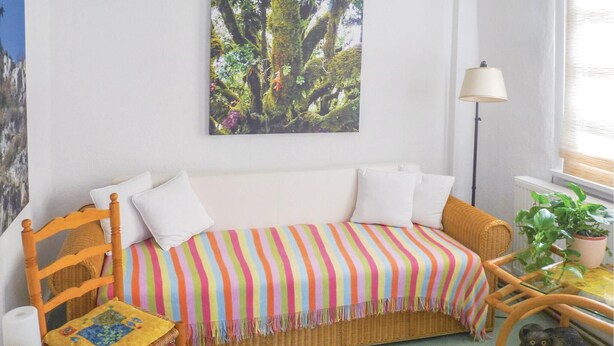 Um quarto acolhedor com um sofá de vime, um cobertor listrado, uma cadeira pequena com almofada, uma luminária e um vaso com planta.