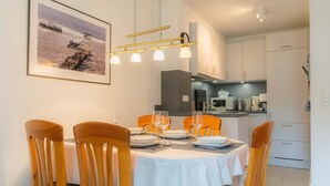 A dining area with a white table set for four, wooden chairs, and a modern pendant light fixture. - House part 19 (ID 032) - House Nordlys (Sankt Peter-Ording)