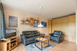Un salon moderne avec un canapé en cuir noir, une table basse et un téléviseur posé sur un meuble en bois. On aperçoit des étagères remplies de divers objets et, au fond, une grande armoire.