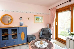 A room with a blue cabinet, a round table with a vase, a polka dot chair, and a large window with a view of greenery.