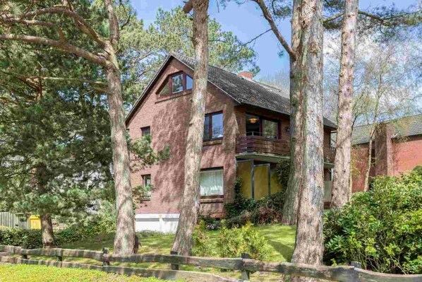 A two-story brick house with a sloped roof, surrounded by mature trees and a well-maintained lawn. - Apartment 2 rear (ID 014) - House Sandpeter (Sankt Peter-Ording)