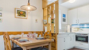 A dining area with a wooden table set for four, a chandelier, and a kitchen with white cabinets and a built-in oven. - House section 8 Greetje (ID 261) (Sankt Peter-Ording)
