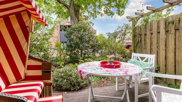 A patio with a striped outdoor sofa, a round table with a floral tablecloth, and a wooden fence. - House section 8 Greetje (ID 261) (Sankt Peter-Ording)