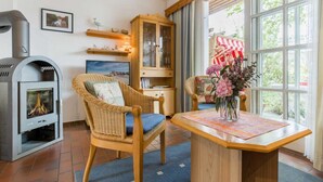 A cozy living room with a fireplace, wicker chairs, a wooden table with a vase of flowers, and a large window with a view of greenery. - House section 8 Greetje (ID 261) (Sankt Peter-Ording)