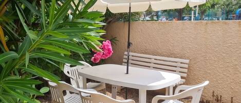Un patio con una mesa y sillas blancas, un banco beige y una sombrilla blanca.