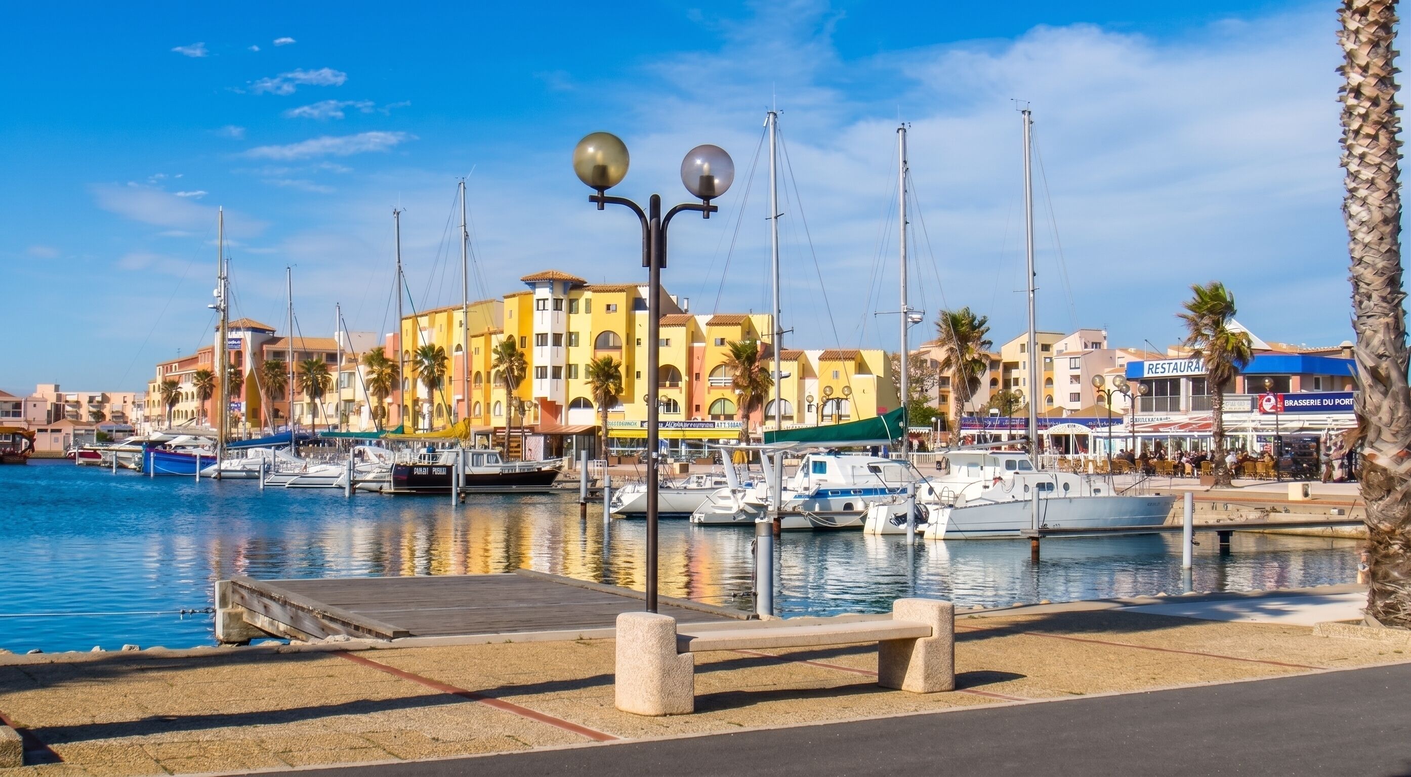 Un puerto deportivo con barcos amarrados, un muelle de madera, edificios con balcones y palmeras.