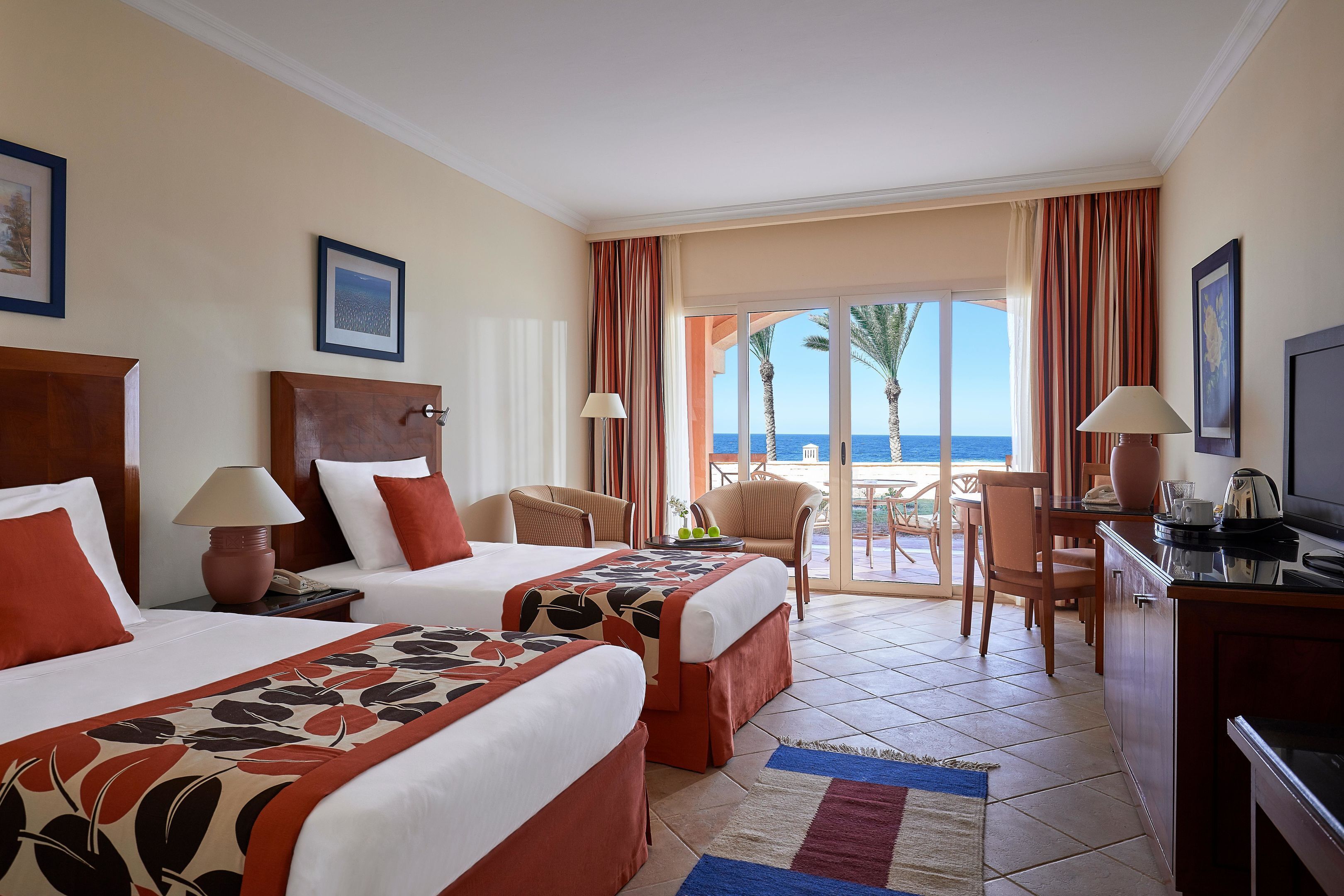 A hotel room with two beds, a desk, a TV, and a balcony with a view of the sea.