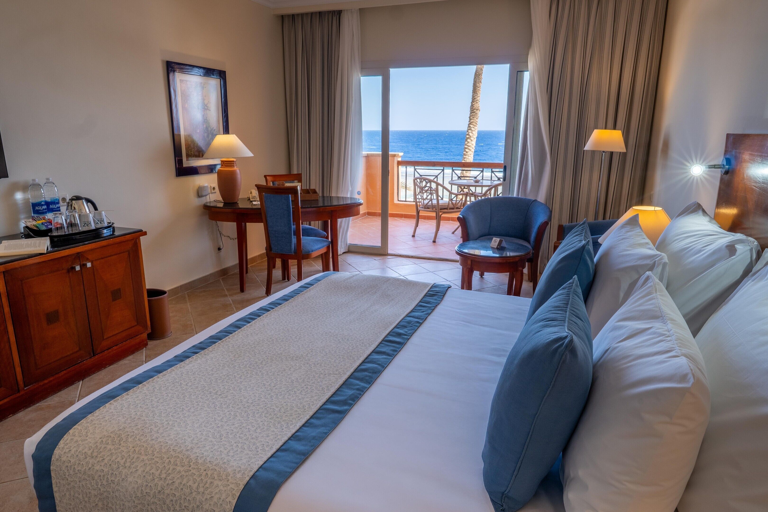 A hotel room with a bed, a desk, a chair, a table, and a view of the sea.