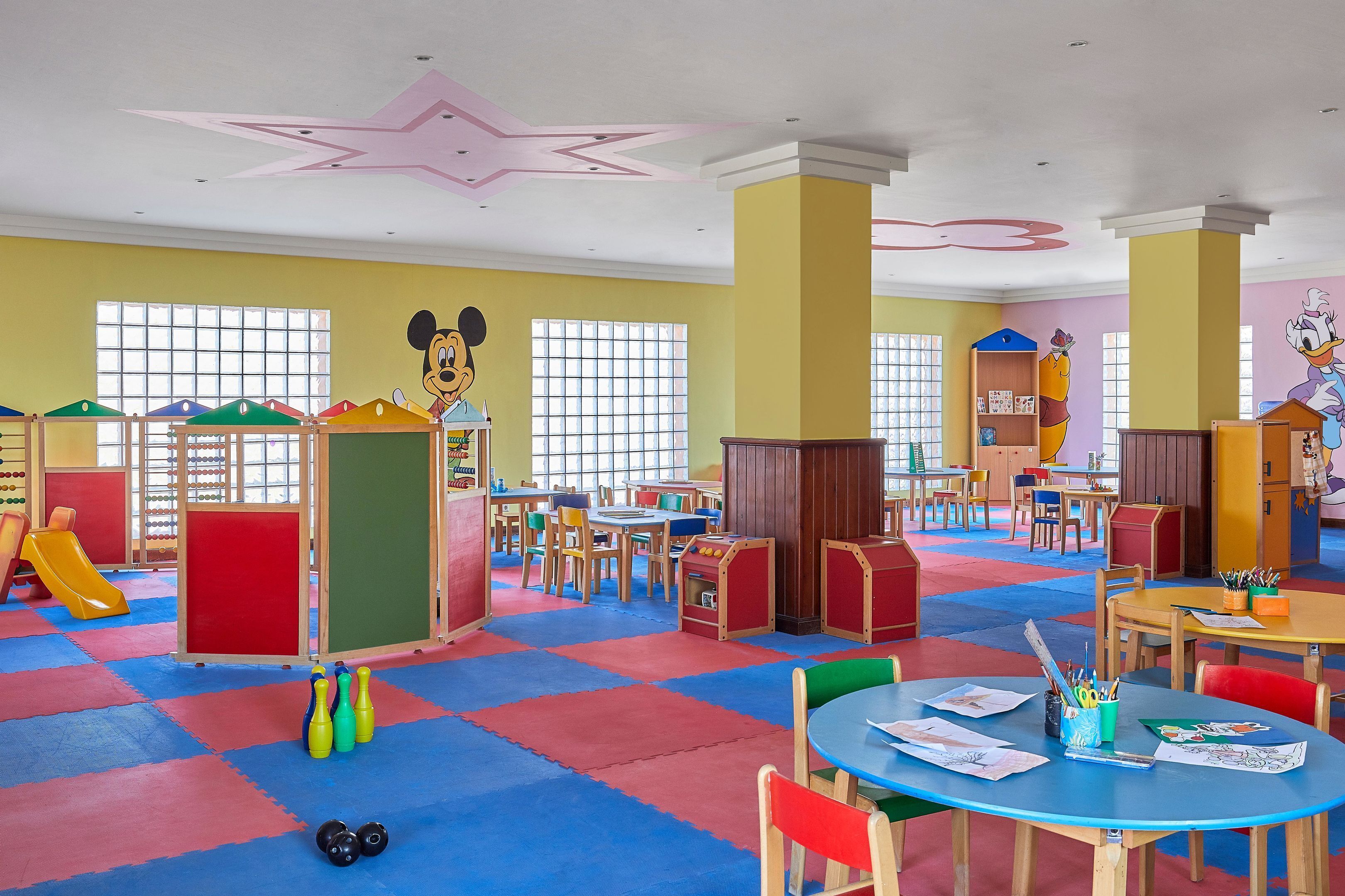 Un área de juegos infantiles con colchonetas de colores, mesas, sillas y decoración de Mickey Mouse.