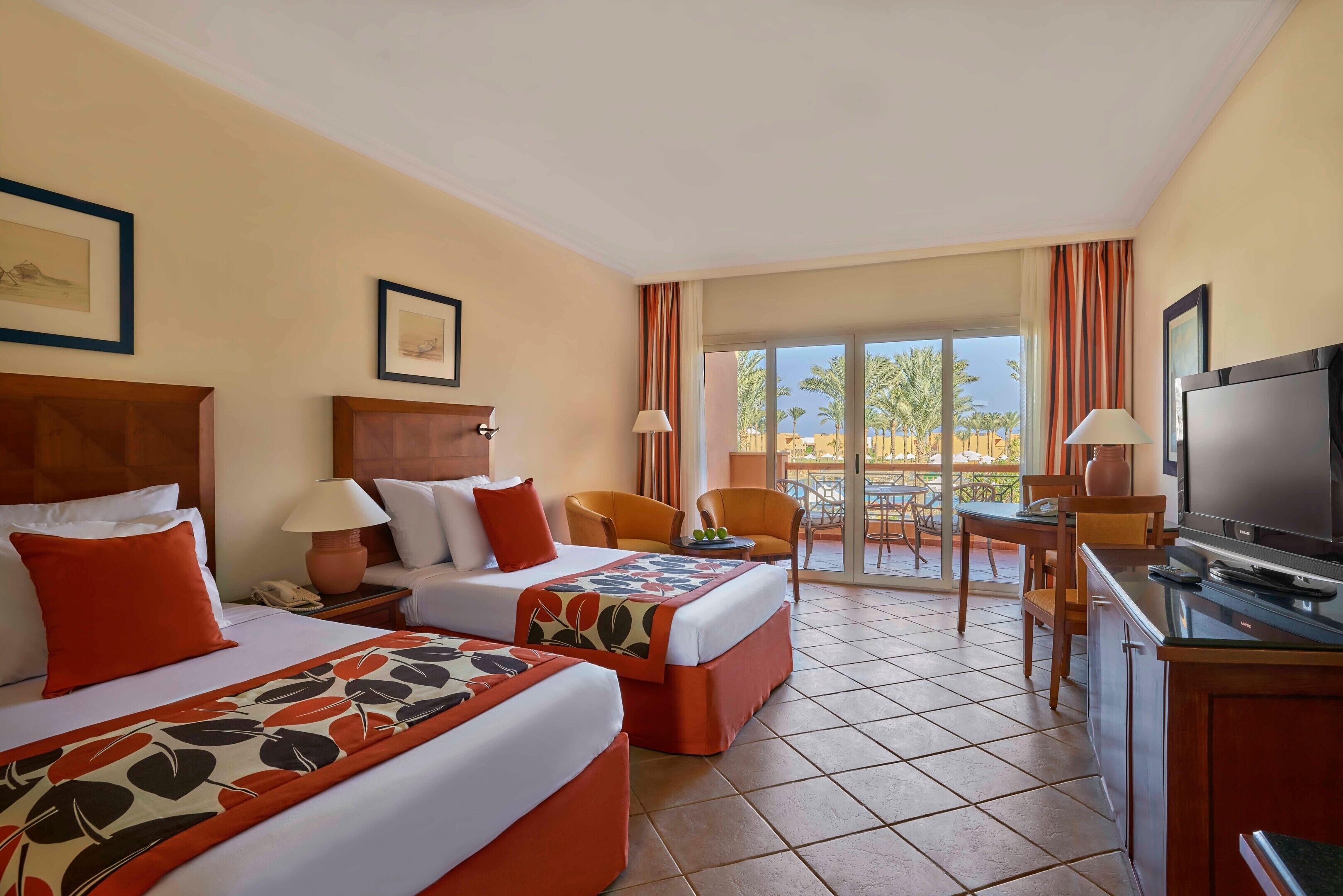 A hotel room with two beds, a TV, a desk, and a balcony with outdoor furniture.