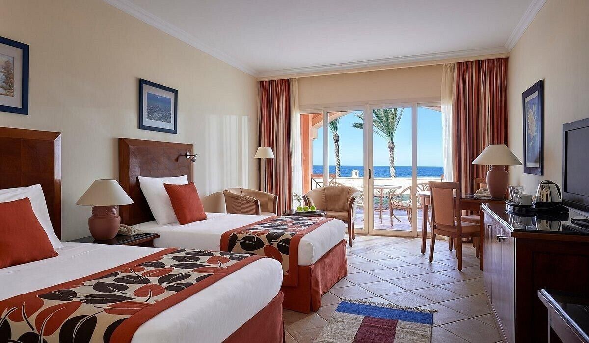 A hotel room with two beds, a desk, a chair, a television, and a view of the ocean.