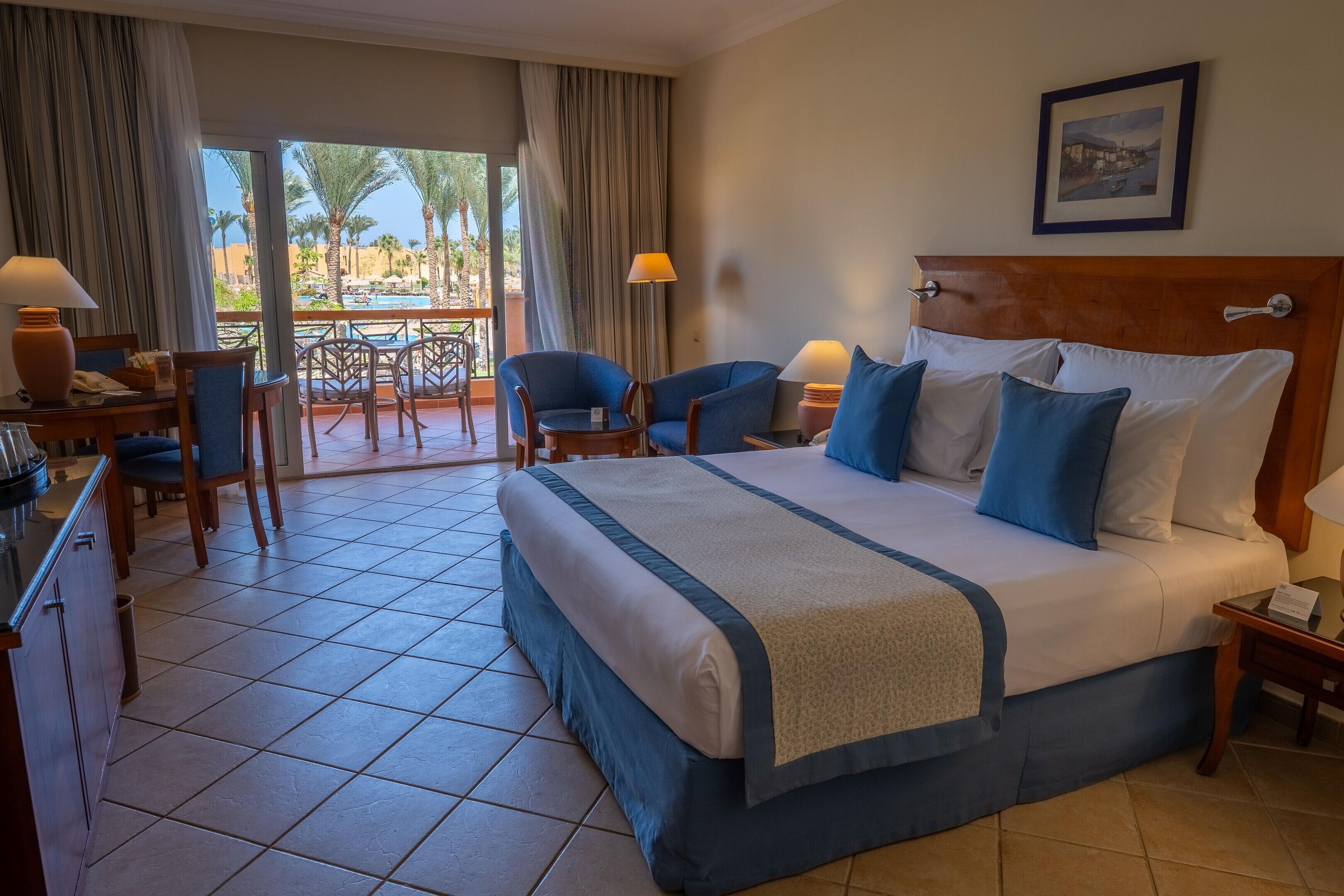 A hotel room with a large bed, two chairs, a desk, and a view of palm trees and a pool.