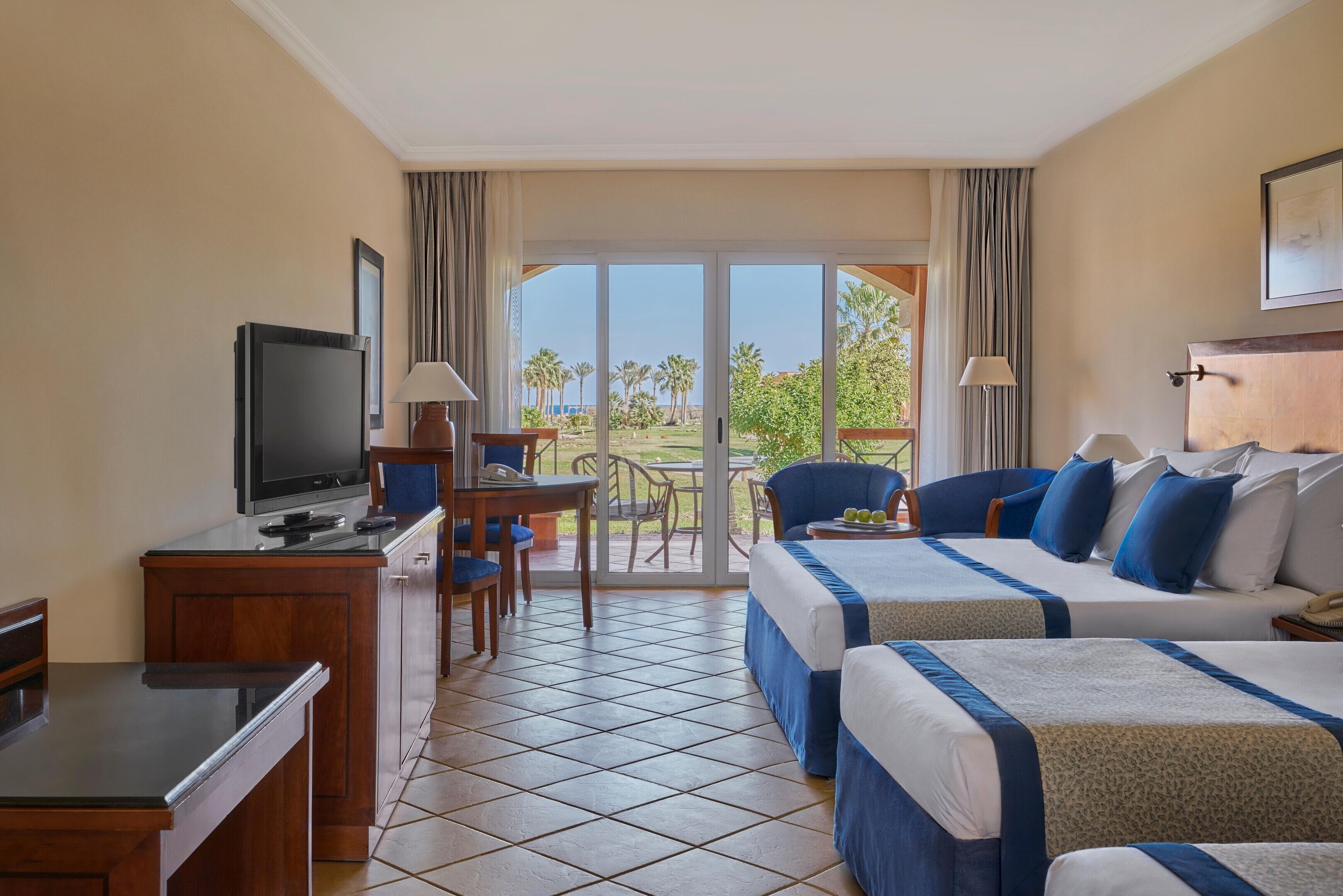A hotel room with a TV, desk, two beds, a chair, and a balcony with a view.