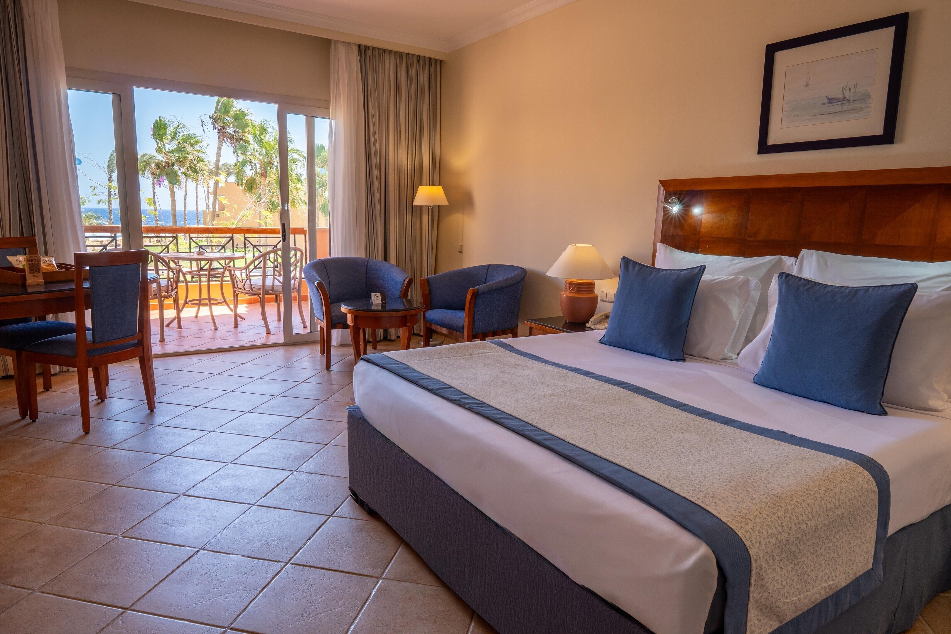 A hotel room with a bed, two chairs, a table, and a view of palm trees and the ocean.