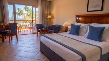 A hotel room with a bed, two chairs, a table, and a view of palm trees and the ocean.