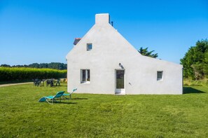 Ein weißes Haus mit einem Giebeldach, einer Tür und Fenstern. Vor dem Haus liegt eine Rasenfläche mit Stühlen und einem Tisch.