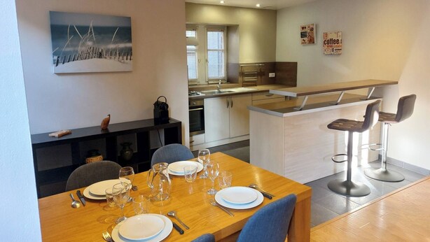 A modern dining area with a wooden table set for four, a kitchen in the background, and a bar area with stools.