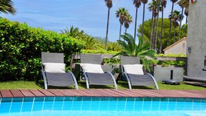A pool area with three lounge chairs, a wooden deck, and palm trees in the background. - Villa Paradisiaque - La HUPPE de SAINT TROPEZ (Saint-Tropez)