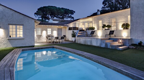 A swimming pool with a wooden deck, a patio with outdoor furniture, and a white building with large windows and a tree in the background. - Villa Paradisiaque - La HUPPE de SAINT TROPEZ (Saint-Tropez)