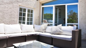 A corner seating area with a wicker sofa and cushions, a glass table, and a large window with a view of trees and a building. - Villa Paradisiaque - La HUPPE de SAINT TROPEZ (Saint-Tropez)