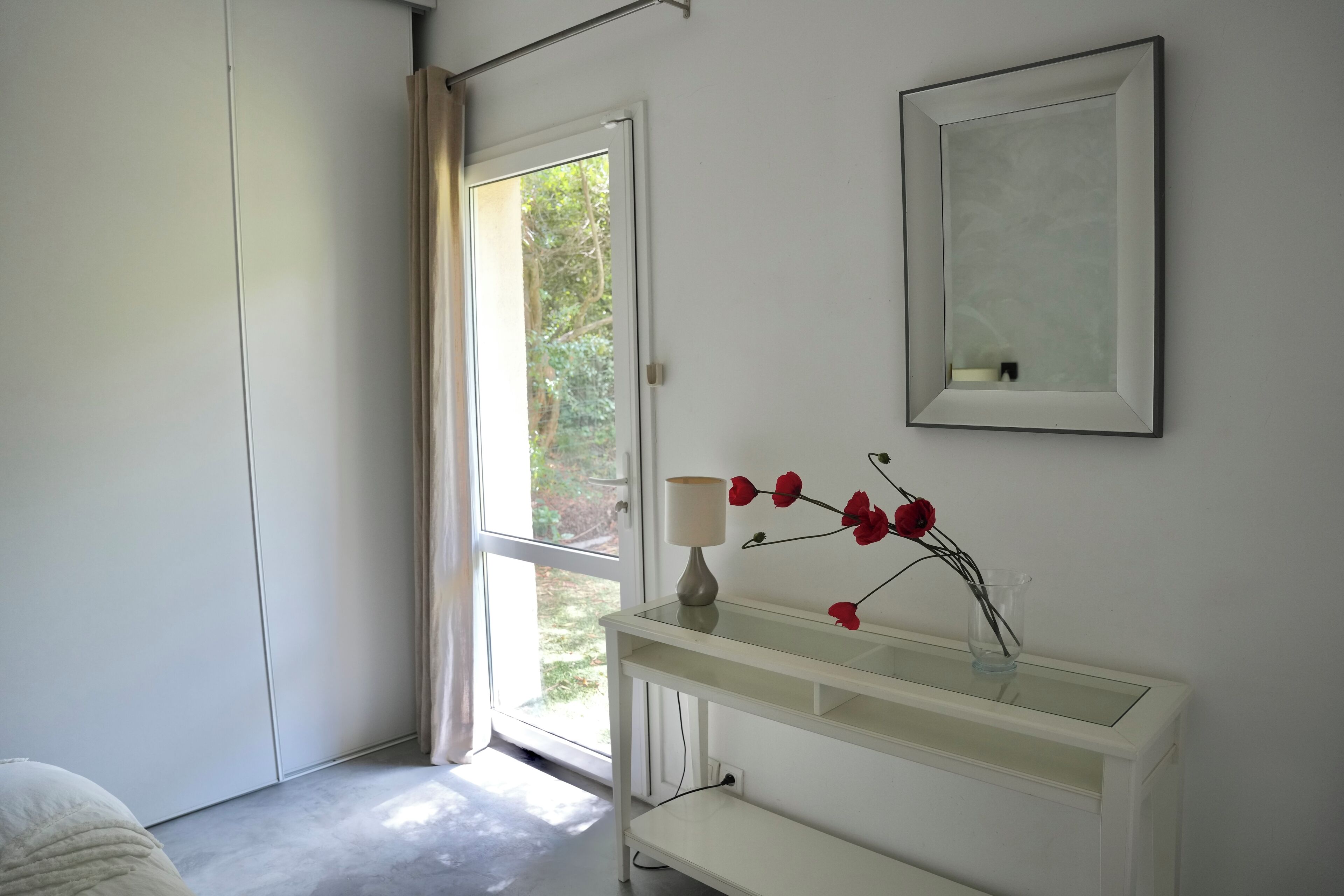 A modern bedroom with a large window, a bedside table with a lamp, and a mirror on the wall.
