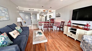 Un salon moderne avec un canapé gris, un plancher en bois, un coin repas avec des chaises rouges et blanches, et une cuisine en arrière-plan.