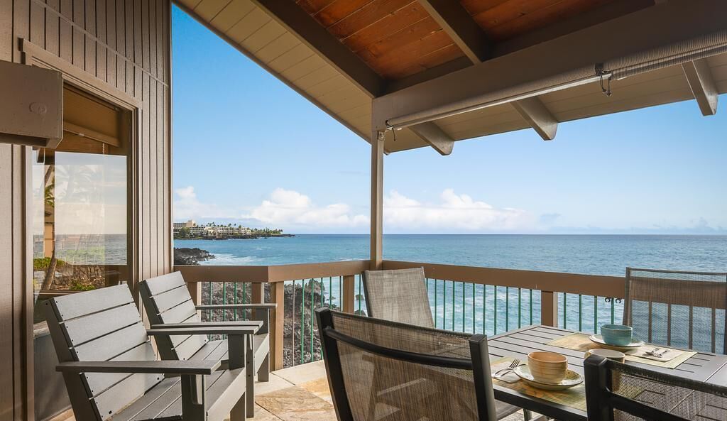A covered outdoor seating area with a table and chairs, overlooking a clear blue sea and a distant shoreline.