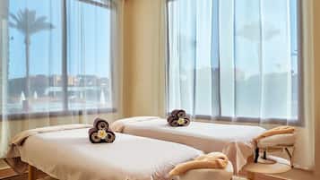 Two single beds with wooden frames and white bedding, set against a backdrop of large windows and dark wood flooring.
