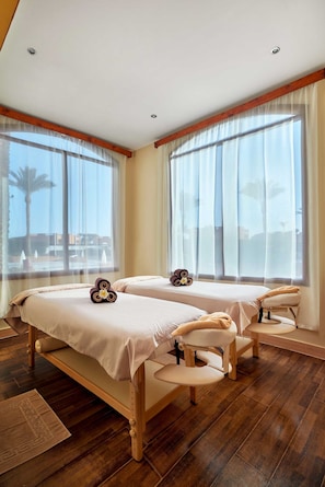 Una habitación con dos camillas de masaje, amplias ventanas con cortinas translúcidas y suelo de madera.