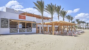 A beachside restaurant with outdoor seating, palm trees, and a clear sky.