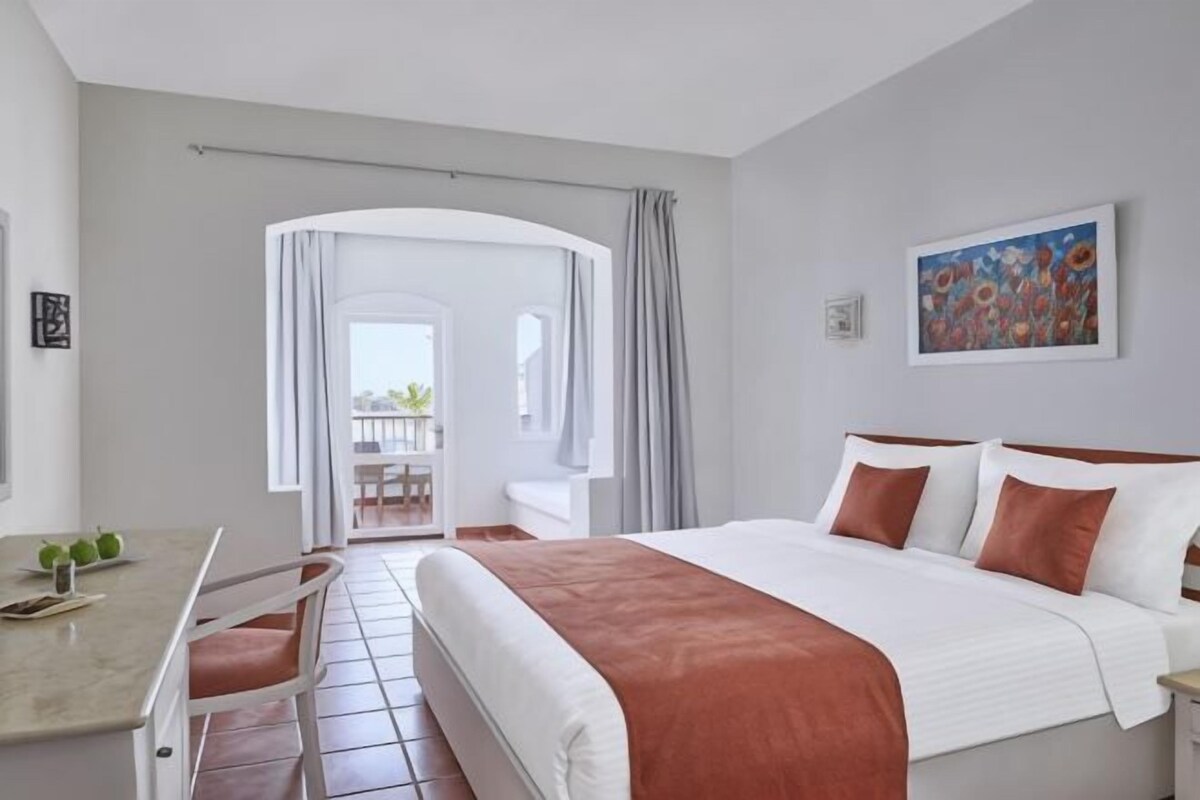 A hotel room with a bed, a desk, a chair, a balcony view, and a painting on the wall.
