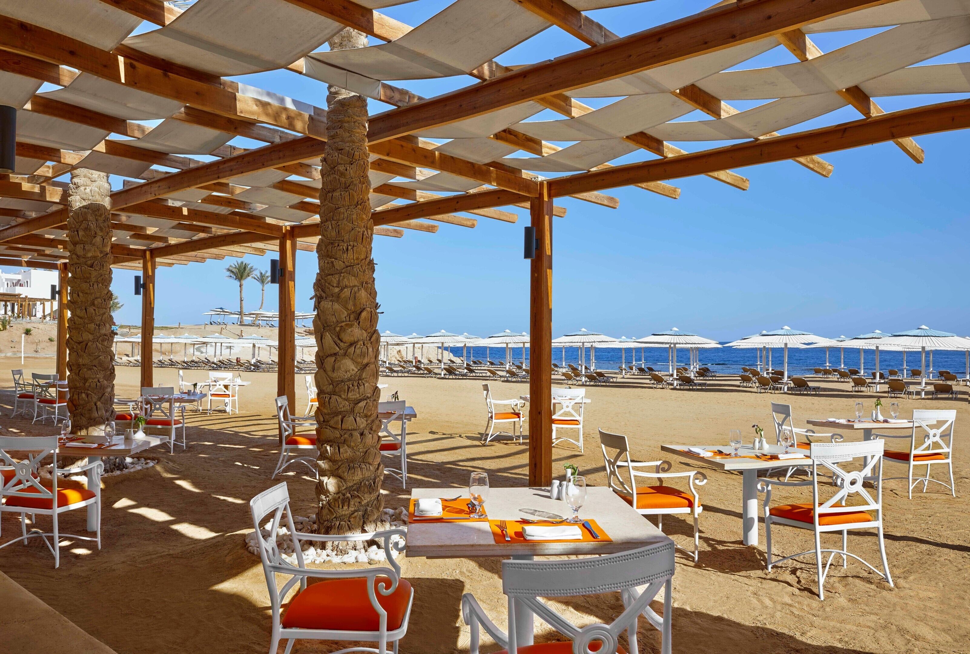 An outdoor dining area with tables and chairs, a pergola structure, and a view of the beach and ocean.