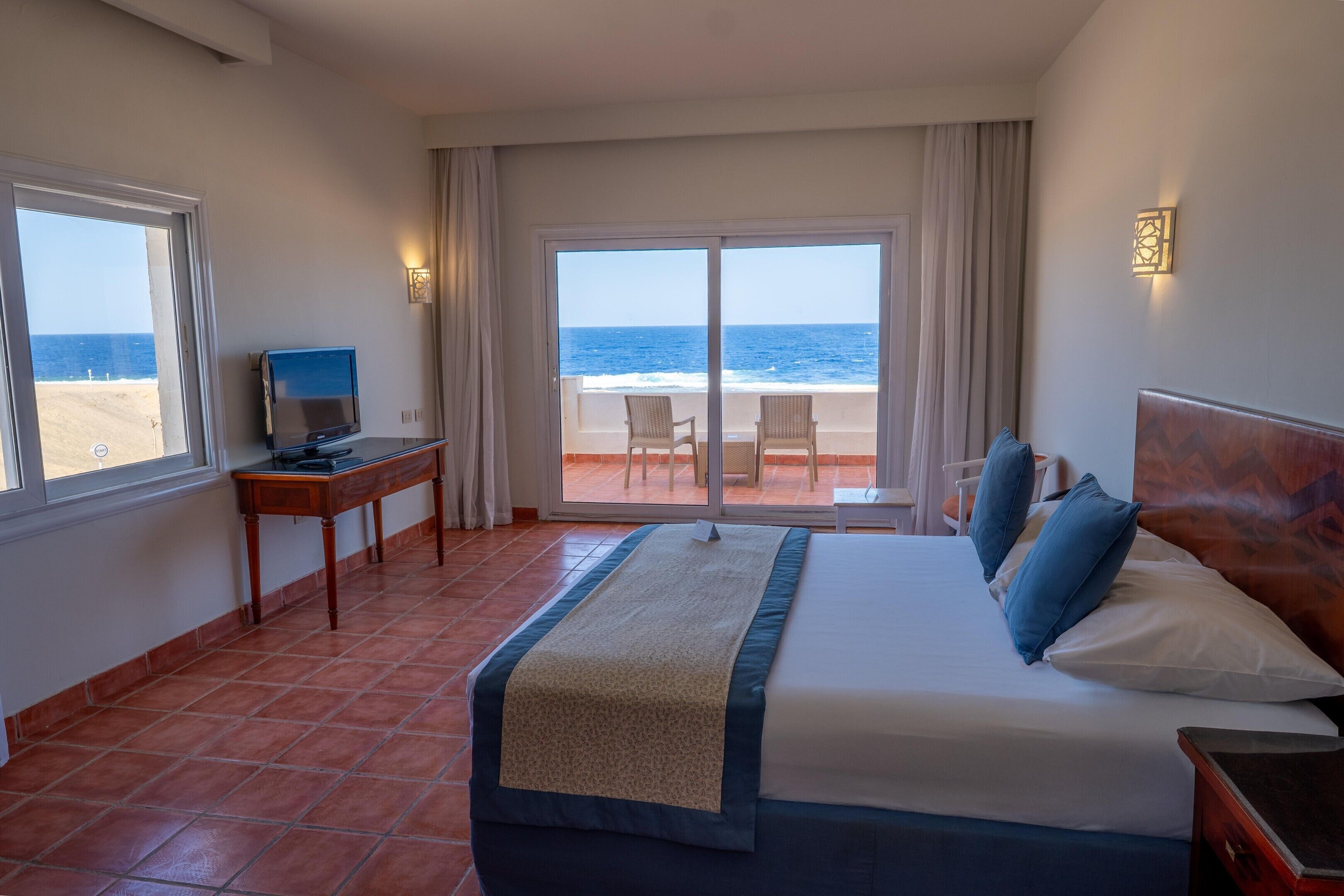 A hotel room with a bed, a desk, a television, and a view of the beach.