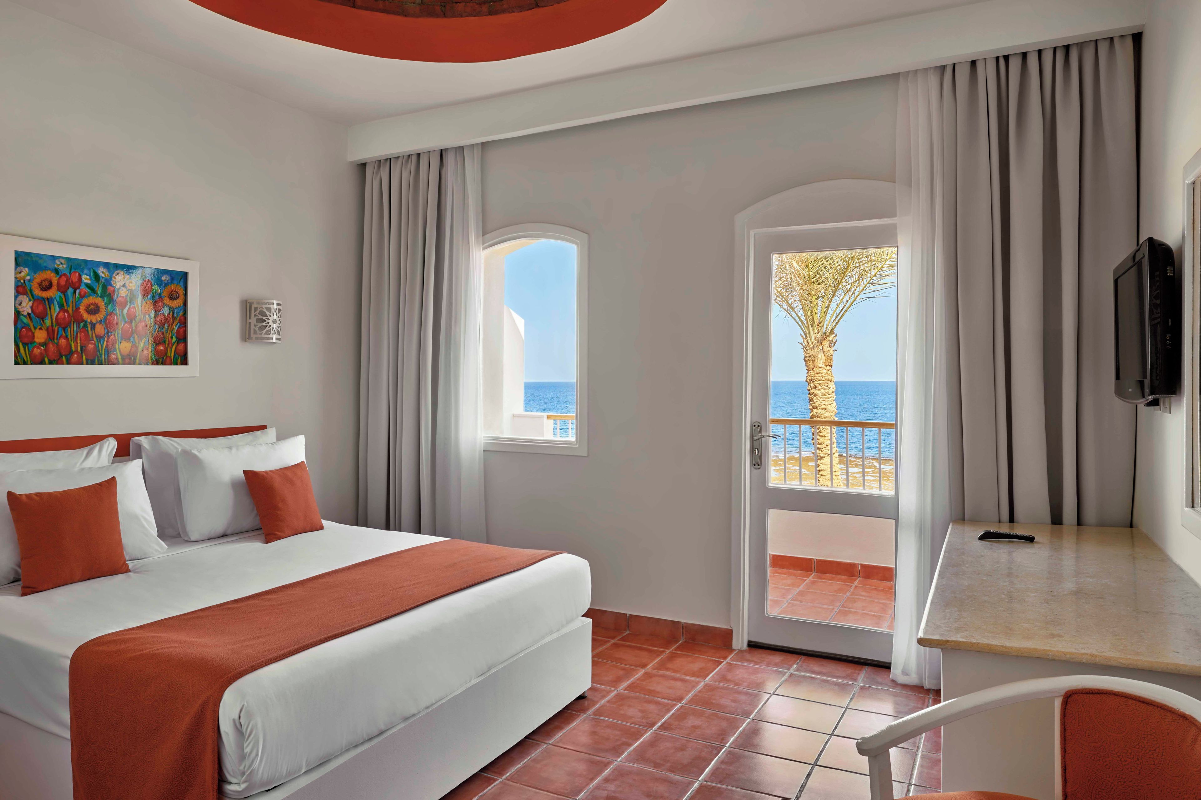 A hotel room with a bed, a desk, a TV, a painting, and a view of the sea.