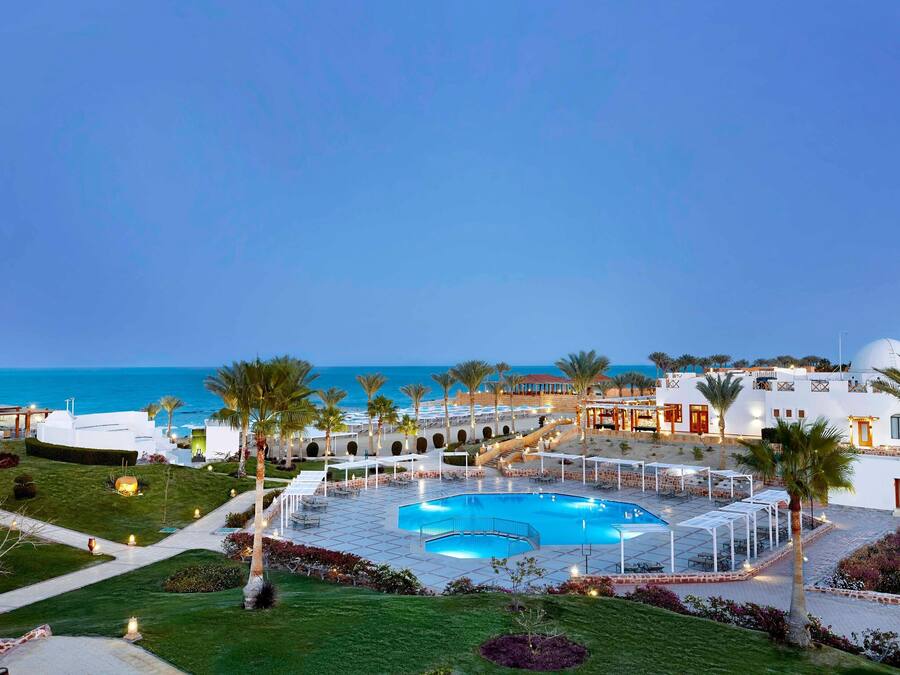 Een resort met een zwembad in het midden, palmbomen en uitzicht op zee.