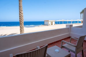 A balcony with wicker chairs and a small table, overlooking a beach with a clear view of the ocean.