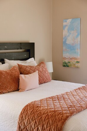 Un lit avec une couverture matelassée, plusieurs oreillers, une tête de lit, une lampe fixée au mur et un tableau représentant un paysage.