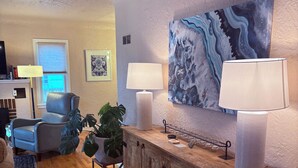A living room with a wooden cabinet, two lamps, a large abstract painting, a blue chair, and a potted plant. - BEST YARD + HOUSE 🏡🤩Hot tub, fire pit, grill, gym, and soaking spa bathtub! (Colorado Springs)