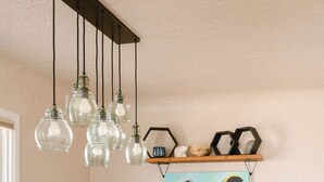 A dining area with a wooden table, chairs, hanging light fixtures, a potted plant, and a wall-mounted shelf with decorative items. - BEST YARD + HOUSE 🏡🤩Hot tub, fire pit, grill, gym, and soaking spa bathtub! (Colorado Springs)
