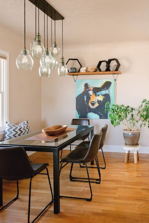 Un coin repas avec une table en bois, des chaises, des luminaires suspendus, une plante en pot et une Ă©tagĂšre murale agrĂ©mentĂ©e dâobjets dĂ©coratifs.