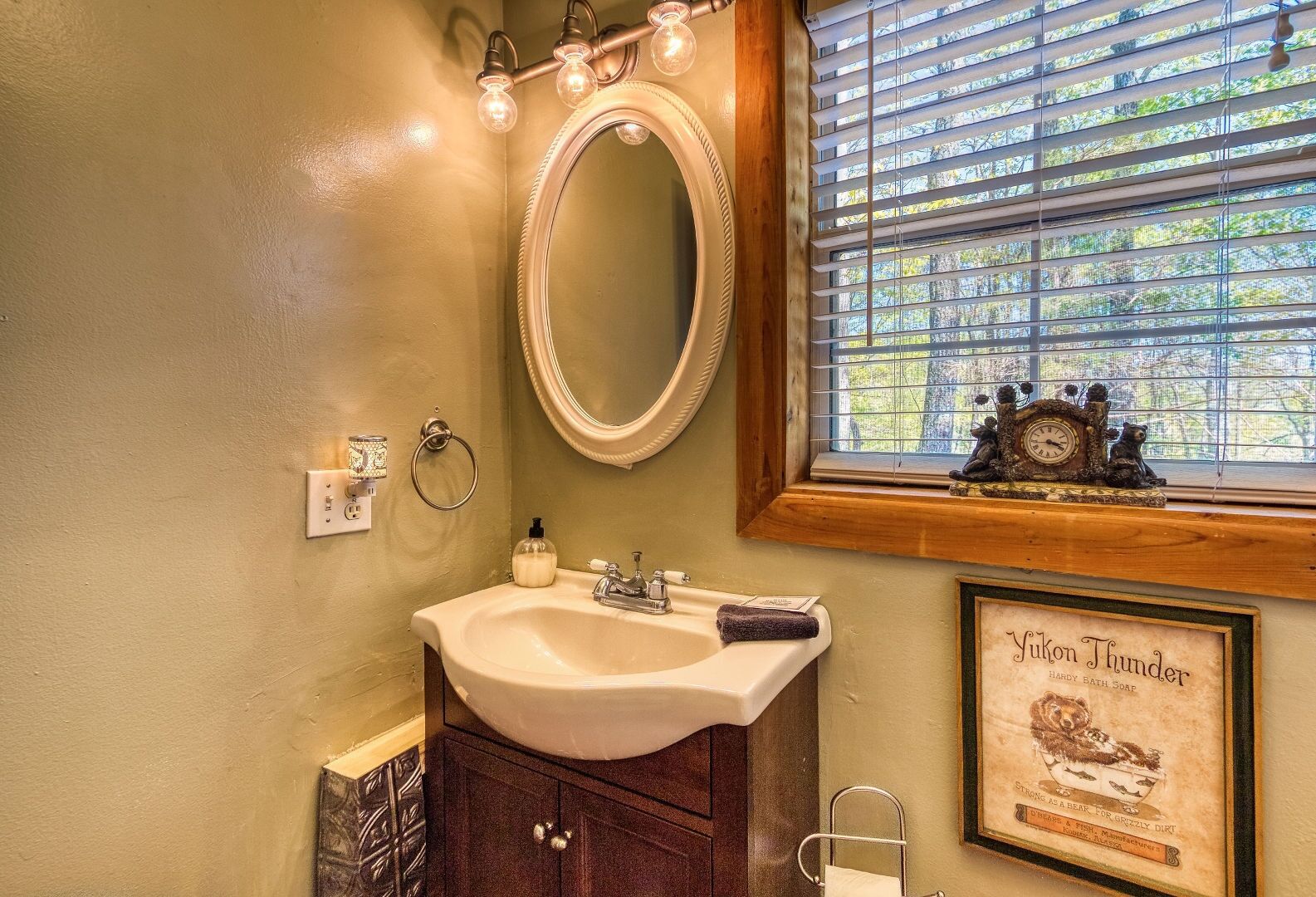 A bathroom with a vanity, a mirror, a window with blinds, and a framed advertisement for Yukon Thunder soap.