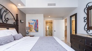 Un dormitorio con cama, cómoda, un cuadro y baño a la vista a través de una puerta abierta.