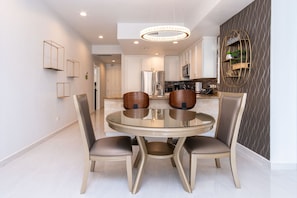 A modern dining area with a round glass table and four chairs, a chandelier, and a kitchen in the background.