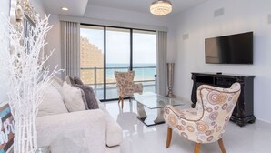 Una sala de estar moderna con un sofá, dos sillones estampados, una mesa de centro de vidrio, un televisor y vistas al mar a través de amplios ventanales.