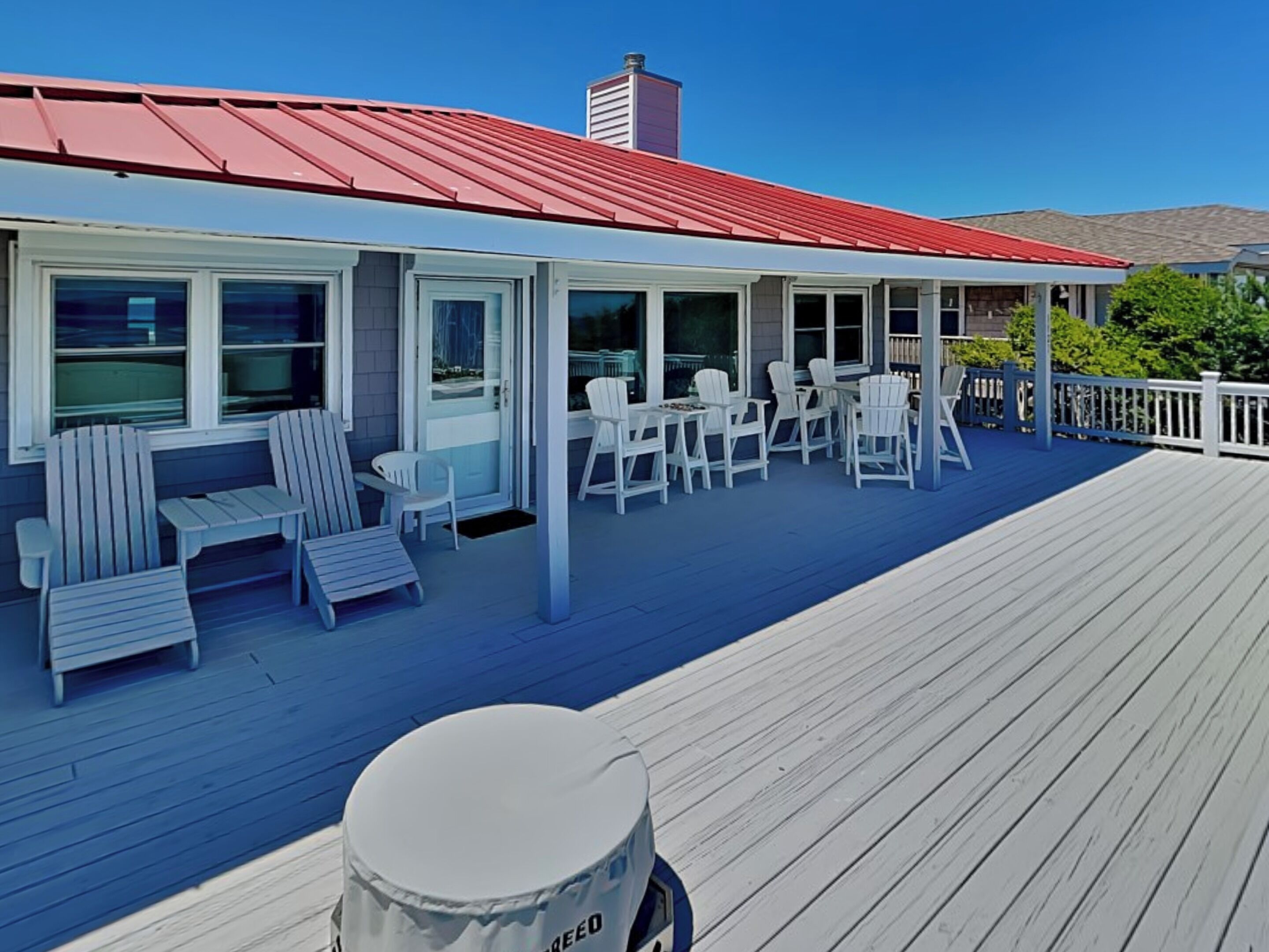 A covered outdoor seating area with white Adirondack chairs and tables, a red roof, and a wooden deck.