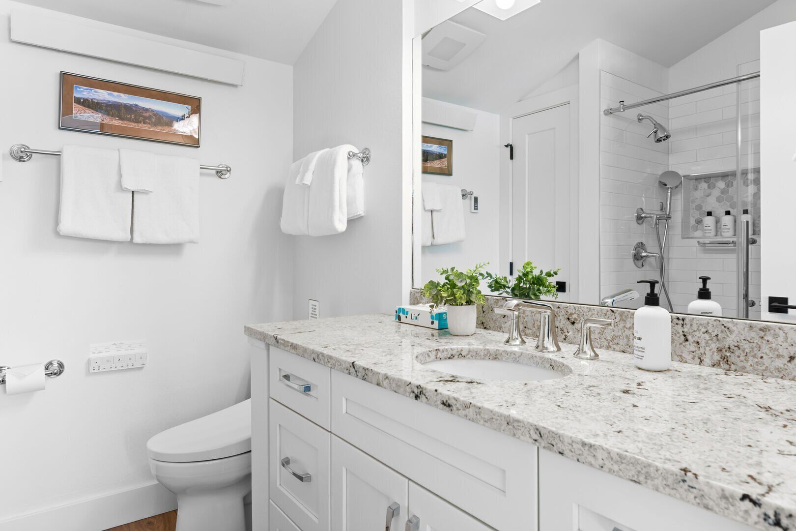 Een moderne badkamer met een wit toilet, een wastafel met een marmeren blad, een douche met een glazen deur en een grote spiegel.