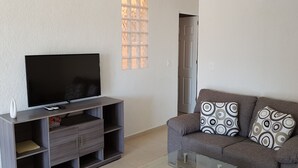 Ein Wohnzimmer mit einem Flachbildfernseher auf einem Holzständer, einem grauen Sofa mit gemusterten Kissen und einem Fenster mit Gittermuster.