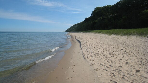 A sandy beach with footprints leading to the water, a clear sky, and a forested hill in the background. - Open for the Holidays! Fireplace, Near XC skiing and Wineries, Good for Groups! (Leland)